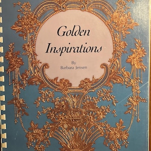 May include: A spiral-bound book titled "Golden Inspirations" by Barbara Jensen. The cover is light blue with a gold floral design surrounding the title. The book has a price tag in the upper right corner.