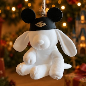 May include: A white plush dog ornament hanging from a jute string. The dog features long floppy ears and wears a black cap with rounded ears and a gold Disney Cruise Line logo on the front. The plush toy is soft and white with stitched details.