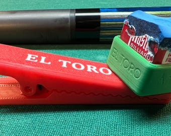 Könnte beinhalten: Ein roter Kunststoffclip mit dem eingeprägten Text EL TORO an der Seite, der einen grünen Kunststoffhalter mit einem blauen Würfel Master-Billardkreide hält. Ein blau-schwarzes Billard-Queue liegt im Hintergrund hinter dem Clip und Halter.