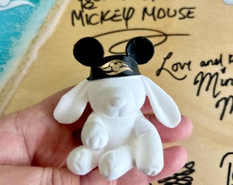 Puede incluir: Una pequeña figura de perro de peluche blanco que lleva una diadema negra con orejas redondas negras y un logotipo dorado de Disney Cruise Line. La figura está colocada frente a una tabla de madera marrón claro con texto escrito a mano Your Pal, Mickey Mouse.