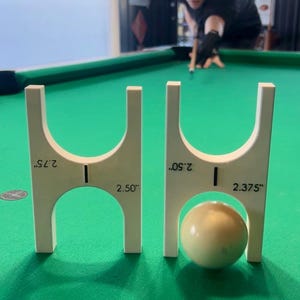 May include: Two white pool ball measuring tools with inch measurements, 2.75", 2.50", and 2.375", are on a green felt pool table. A white pool ball is between the tools. A person is in the background, aiming a pool cue.