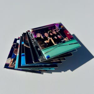 Könnte beinhalten: Ein Stapel glänzender, rechteckiger Magnete mit Gruppenfotos. Der oberste Magnet zeigt Personen an einem Billardtisch. Die Magnete sind auf einer weißen Oberfläche angeordnet und werfen einen Schatten.