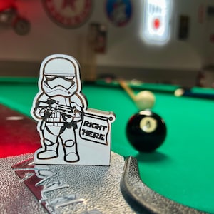 Puede incluir: Una figurita de madera blanca y negra de un Stormtrooper de Star Wars sosteniendo un cartel que dice "Right Here". La figurita está sobre una mesa de billar de fieltro verde.