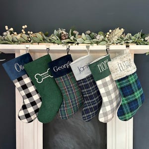 May include: Six personalized Christmas stockings hanging in a row. The stockings feature various patterns including black and white buffalo check, solid green with a bone icon, green plaid, navy blue plaid, gray buffalo check, and blue plaid.