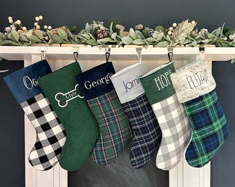 Op de afbeelding: Zes gepersonaliseerde kerstsokken hangen op een rij. De sokken hebben verschillende patronen, waaronder zwart-wit geblokt, effen groen met een bot-icoon, groen ruitpatroon, marineblauw ruitpatroon, grijs geblokt en blauw ruitpatroon.