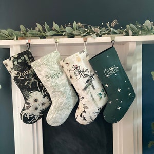 May include: Four personalized Christmas stockings hanging in a row. The first is black with white floral patterns and the name Quinn. The second is light green with pine needle prints and the name Harper. The third is white with floral and pine designs and the name Riley. The fourth is dark green with white star shapes and the name Charlie.