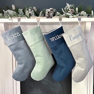 Könnte beinhalten: Vier personalisierte Weihnachtsstrümpfe hängen an einem weißen Kaminsims. Die Strümpfe sind aus Samt und in den Farben Blau, Grün und Grau gehalten. Die Namen Luca, Olivia, Willow und Emma sind in weißer Schrift auf die Strümpfe gestickt.