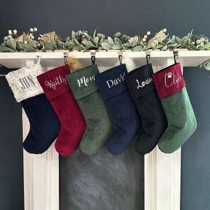 May include: Six hanging Christmas stockings in various colors including navy, burgundy, forest green, and black. Each stocking features a personalized name embroidered in white or black script, with one stocking displaying a paw print icon.