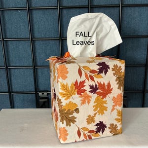 May include: A square fabric tissue box cover featuring a pattern of autumn leaves in shades of orange, yellow, brown, and purple. A white tissue protrudes from the top, printed with the black text FALL Leaves. An orange ribbon is tied at the top.