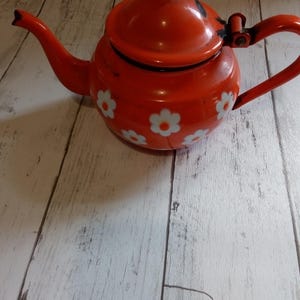 May include: A red enamel teapot with a lid and handle. The teapot is decorated with white daisy flower patterns. The teapot is sitting on a light-coloured wooden surface.