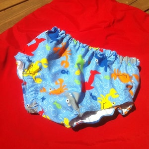 May include: Blue and white swim diaper with a colorful sea creature print. The print features crabs, fish, and bubbles.
