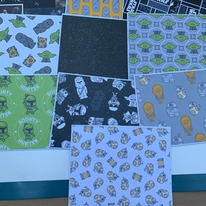 May include: A collection of Star Wars themed scrapbook paper featuring various characters and designs. The paper includes a black and white pattern with the words "Star Wars", a yellow pattern with a repeating image of a character, a green pattern with a repeating image of Yoda, a gray pattern with a repeating image of a droid, and a white pattern with a repeating image of a droid.
