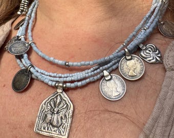 May include: A layered necklace with light blue beads and silver charms. The charms include a pendant with an intricate design, a coin with a profile of a woman, and a heart-shaped charm.