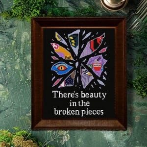 May include: A framed artwork with a dark brown wooden frame. The artwork features a colorful, abstract design on a black background, with the text "There's beauty in the broken pieces". The artwork is displayed on a green marble surface.