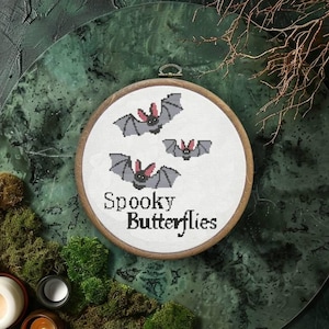May include: A white embroidery hoop with a cross-stitch design of three bats flying. The text "Spooky Butterflies" is stitched below the bats.