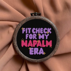 May include: A round, cross-stitched embroidery hoop with the text "FIT CHECK FOR MY NAPALM ERA" in purple and pink on a black background. The hoop has a dark wood-grain effect and is set against a peach-colored fabric.