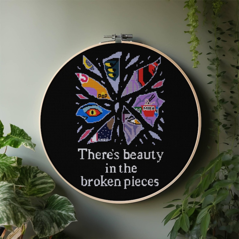 May include: A black cross-stitch embroidery hoop with a design of fragmented shapes in various colors, including red, yellow, and purple. The text "There's beauty in the broken pieces" is stitched below the design. The hoop is set against a neutral background.