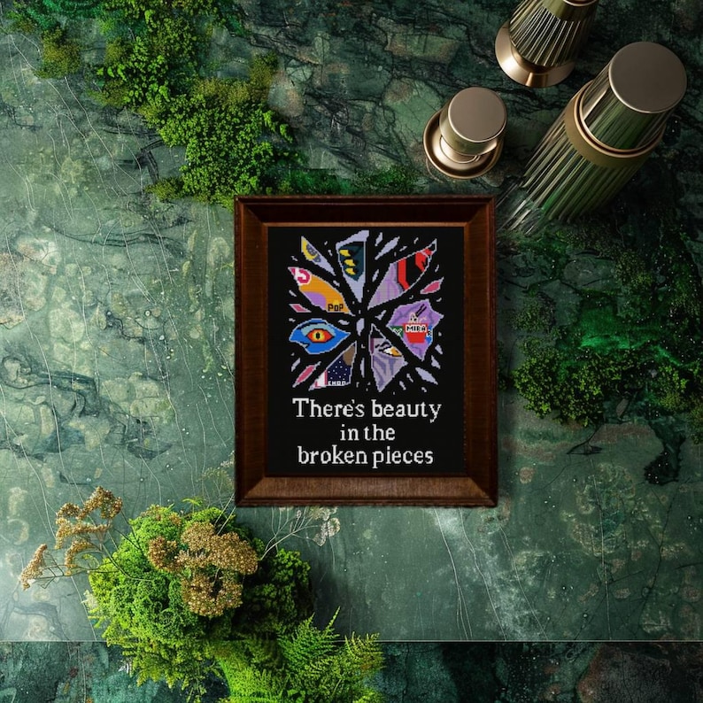 May include: A framed artwork with a dark brown wooden frame. The artwork features a colorful, abstract design on a black background, with the text "There's beauty in the broken pieces". The artwork is displayed on a green marble surface.