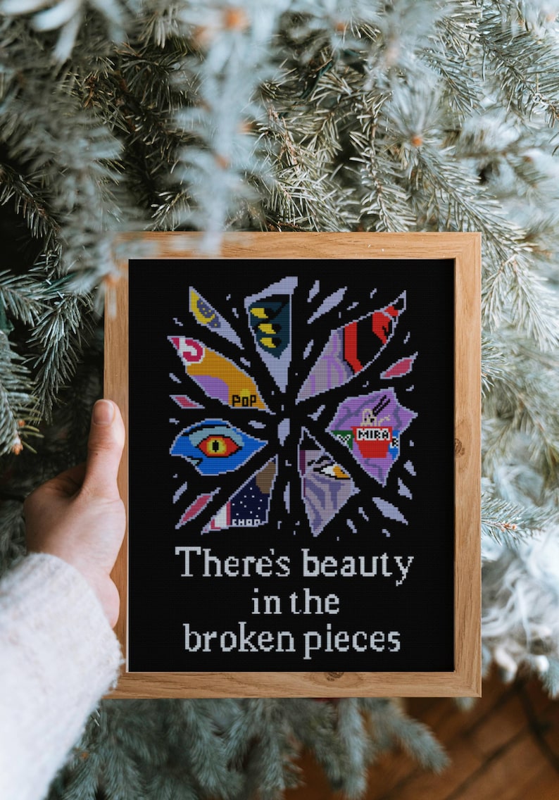 May include: A framed cross-stitch artwork on black fabric, featuring a fragmented design with colorful geometric shapes and the text "There's beauty in the broken pieces." The frame is made of light-colored wood.