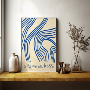 Könnte beinhalten: Ein gerahmter Druck mit cremefarbenem Hintergrund und blauen abstrakten Linien. Der Text "ici la vie est belle" ist unten auf dem Druck gedruckt.