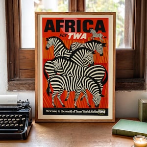 Peut inclure: Une affiche de voyage vintage encadrée pour l'Afrique, avec un fond rouge vif et le mot "AFRICA" en noir. L'affiche présente un groupe de zèbres aux rayures noires et blanches. Le texte "FLY TWA" est également visible.