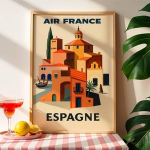 May include: A framed vintage travel poster for Air France, featuring a stylized illustration of a Spanish village with orange and yellow buildings. The poster includes the text "AIR FRANCE" and "ESPAGNE". A cocktail and fruit sit in front of the poster.