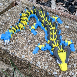 May include: A 3D-printed articulated dragon toy in yellow, blue, black, and white. The dragon has a segmented body, four legs, and a detailed head with pointed ears. The toy is lying on a rough, gray stone surface.