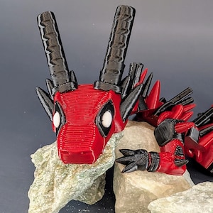 May include: A 3D printed red and black dragon figurine with a Deadpool-inspired design. The dragon is lying on its belly with its head resting on a rock. The dragon has black horns and spikes, and its eyes are white with black pupils.