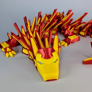 May include: A 3D-printed articulated dragon toy in red and yellow. The dragon's head is yellow with red accents, resembling a superhero mask. The body and limbs are red with yellow spikes and details. The toy is designed to be flexible and poseable.