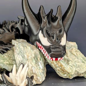 May include: A black 3D printed dragon figurine with red and white details. The dragon is posed with its head resting on a rock.
