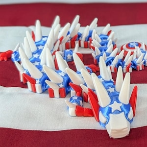 May include: A 3D printed dragon made of white plastic with red, white, and blue stars and stripes. The dragon is curled up in a circle with its head resting on its tail. The dragon is on a red, white, and blue striped background.