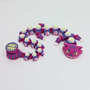 May include: A collection of whimsical, handmade polymer clay figures in shades of pink, blue, and white. The set includes a clock, a segmented caterpillar-like creature, and a Cheshire Cat head, all with intricate details and a playful aesthetic.
