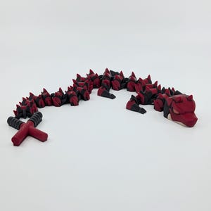 May include: A red and black articulated dragon toy. The dragon has a segmented body with red spikes and black accents. The head and tail are also red and black. The toy is on a white surface.