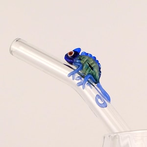 May include: A clear glass drinking straw with a blue and green chameleon figurine attached. The chameleon has red eyes and is perched on the straw. The straw is angled towards a clear glass.