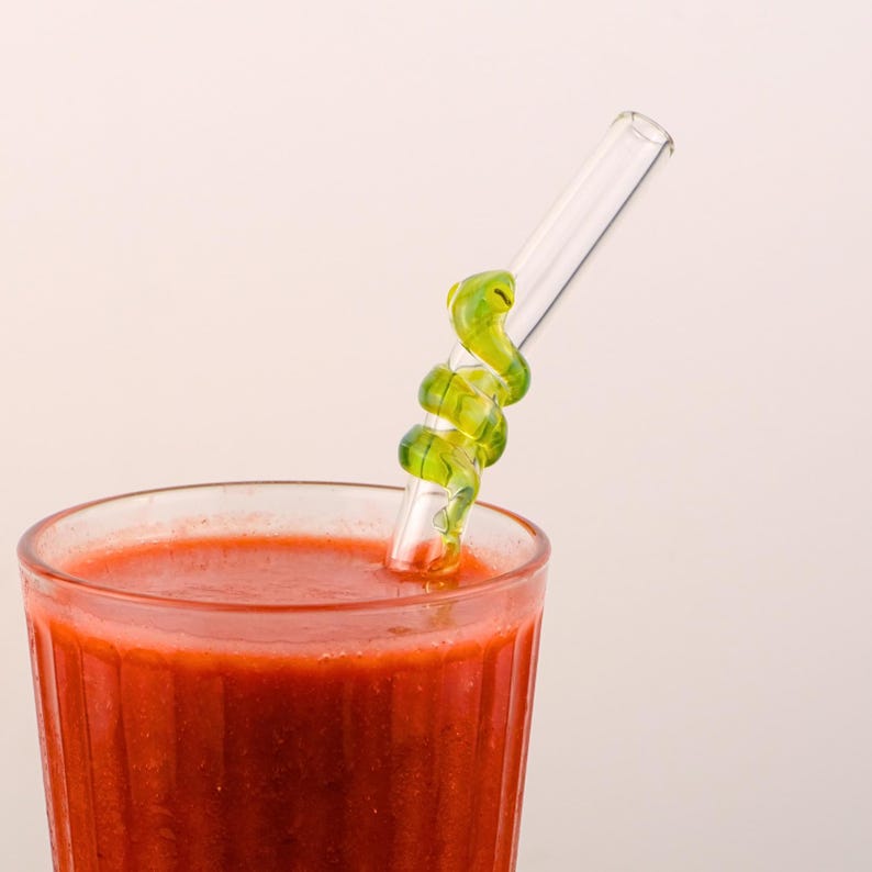 Pu&ograve; includere: Una cannuccia di vetro trasparente con un design a spirale verde &egrave; inserita in un bicchiere di liquido rosso. L'elemento decorativo della cannuccia aggiunge un tocco unico alla bevanda. Il bicchiere ha un design a coste.