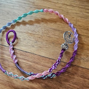 Puede incluir: Un collar de macramé hecho a mano en tonos morado, rosa y turquesa. El collar presenta un diseño en espiral y un dije plateado con un motivo de árbol de la vida. El cierre de bucle es de color morado oscuro.