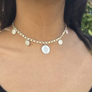 May include: Silver-toned necklace with a chain-link design and multiple dangling charms. The charms include small round pendants and a larger circular pendant with an intricate design. The necklace is worn around the neck.