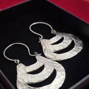May include: A pair of silver-colored earrings with a hammered texture and a unique design featuring two curved cutouts. The earrings are displayed in a black box, with a red and patterned background.