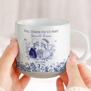 May include: White ceramic mug with a blue floral design and the text "Till Death Do Us Part James & Emma EST.2016". The mug features a skeleton couple illustration. The mug is held by a person.