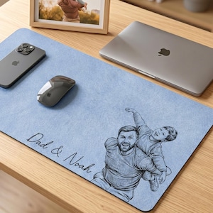 Op de afbeelding: Een lichtblauwe bureaumat met een schets van een vader en zoon, de woorden "Dad & Noah" zijn cursief geschreven. Een ingelijste foto, een laptop, een smartphone en een muis liggen op het bureau.