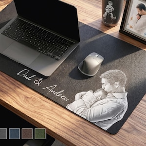 Könnte beinhalten: Eine schwarze Schreibtischunterlage mit den Worten "Dad & Andrew" in weißer Schrift. Die Unterlage zeigt ein graviertes Bild eines Vaters, der ein Baby hält. Ein Laptop, eine Maus und ein gerahmtes Foto befinden sich auf dem Holzschreibtisch.