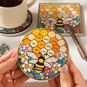 Peut inclure: Sous-verres en céramique avec un motif d'abeille et le mot "GRAND-MÈRE". Les sous-verres présentent un motif en nid d'abeille avec des noms et des fleurs. Une tasse de café repose sur un sous-verre assorti. Un stylo est également visible.