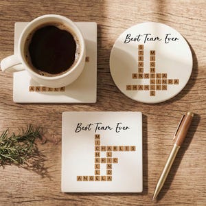May include: Three white ceramic coasters with the text "Best Team Ever" and names spelled out in wooden letter tiles. One coaster holds a white coffee cup filled with dark coffee. A pen rests on the wooden surface.