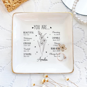 Peut inclure: Un vide-poche carré en céramique blanche avec un bord doré. Le plat présente les mots "YOU ARE..." et des mots d'inspiration avec des versets bibliques. Une bague et un accessoire en forme de fleur reposent sur le plat.