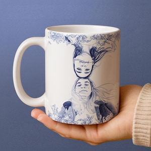 May include: White ceramic mug featuring a blue illustration of two women's faces, one inverted above the other. The design includes floral accents and is set against a blue background. The mug has a curved handle.