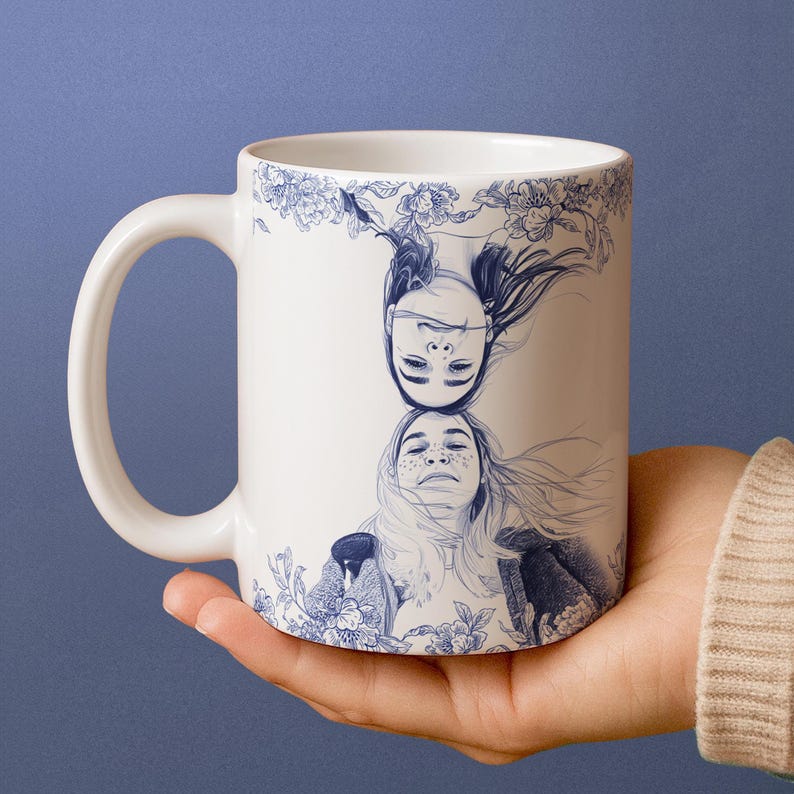 May include: White ceramic mug featuring a blue illustration of two women's faces, one inverted above the other. The design includes floral accents and is set against a blue background. The mug has a curved handle.