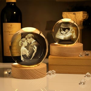 May include: Two illuminated crystal spheres on wooden bases. One sphere displays a couple embracing, the other an ultrasound image. The spheres are lit from below, creating a warm glow. Decorative crystals are scattered around.