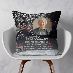 Peut inclure: Un coussin d&eacute;coratif avec une photo d'une femme, d'un arbre et d'oiseaux rouges. Le coussin porte l'inscription "A HUG FROM Heaven" et un message sur l'amour et les c&acirc;lins. Le coussin est sur une chaise blanche.