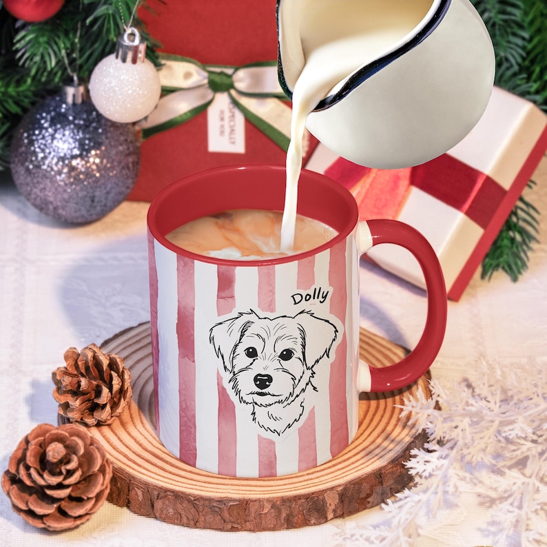 May include: A red and white striped mug with a dog illustration and the name "Dolly" is filled with a creamy liquid. The mug sits on a wooden coaster, with pinecones and Christmas decorations in the background. A white pitcher pours liquid into the mug.