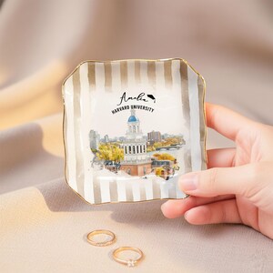 May include: Square ceramic trinket dish with a gold rim and vertical beige stripes. The center features a watercolor illustration of a building with a blue dome, with the text "Amelia Harvard University". Two gold rings are in the foreground.